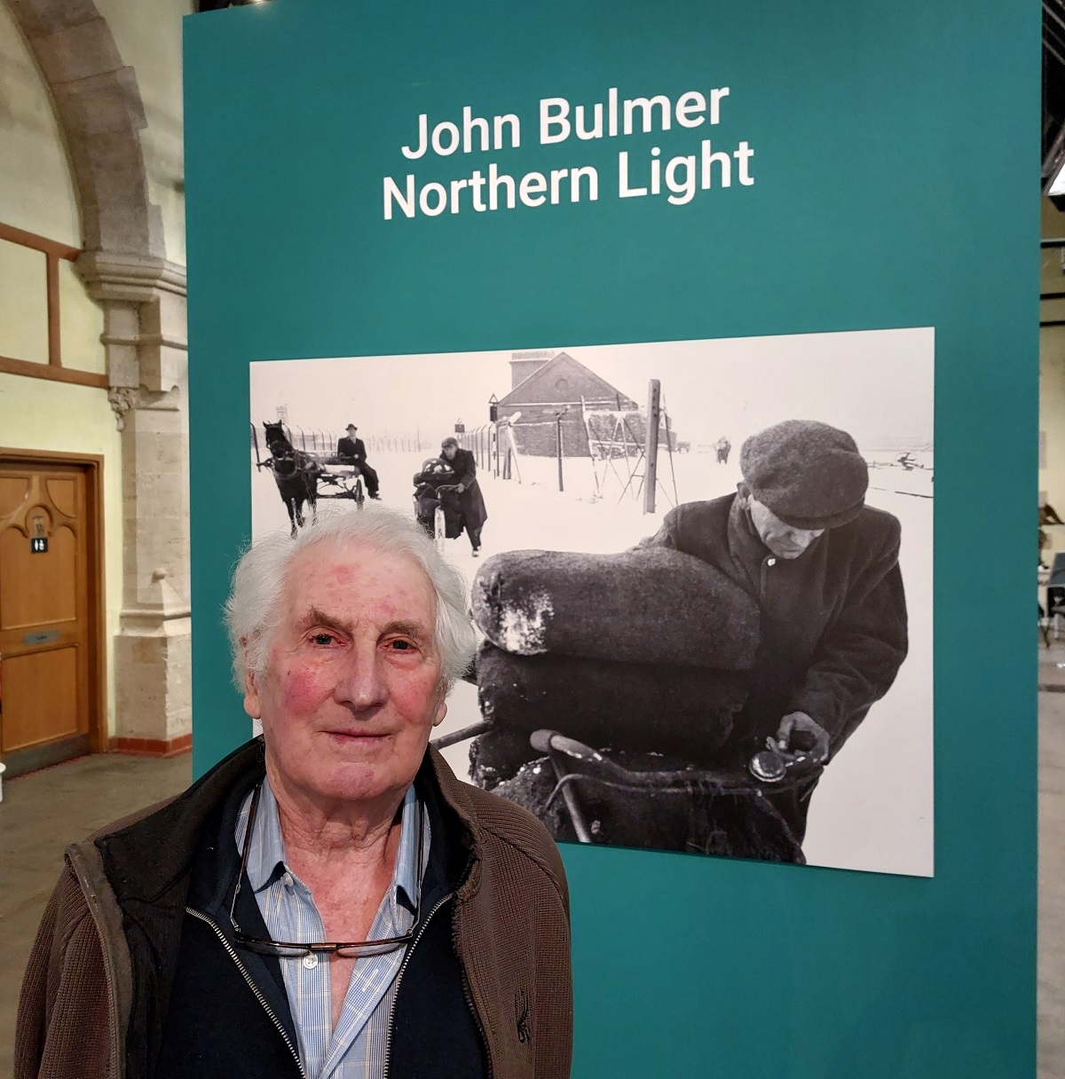 Photographer stands outside the John Bulmer Northern Light Exhibition with a black and white image behind him
